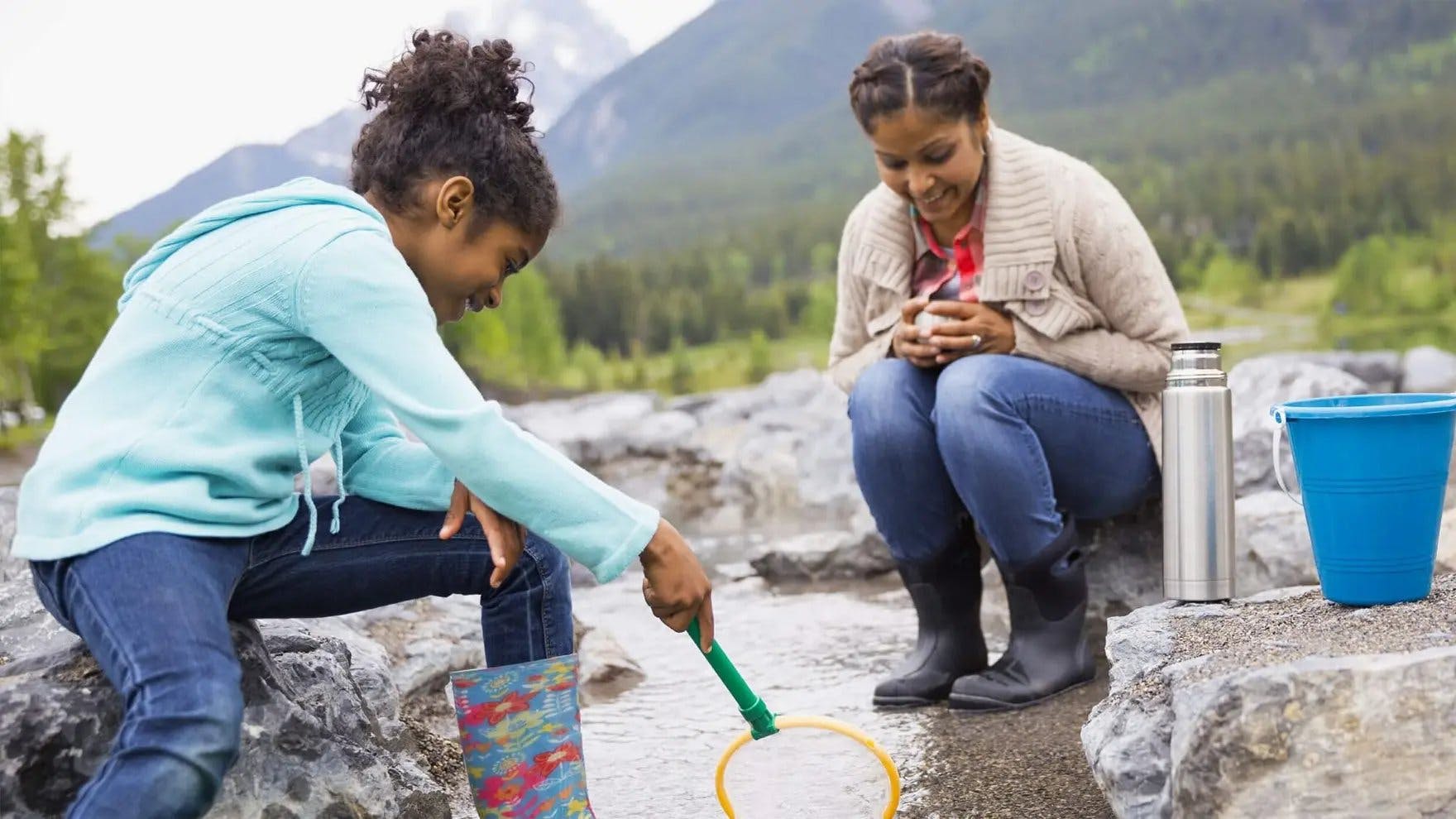 Mulher e criança usam suéteres e botas de chuva, e uma rede para pegar insetos em um riacho raso