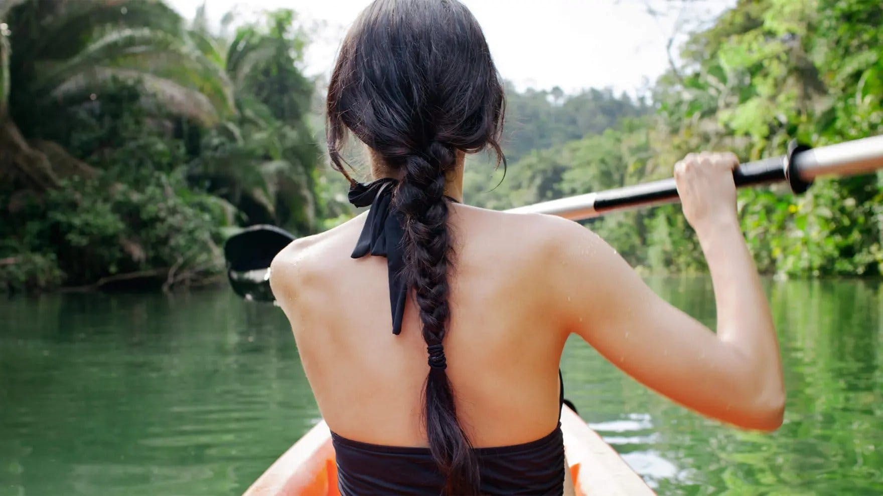 Mulher com cabelo preto longo com uma trança andando de caiaque em um rio com as costas viradas para a câmera